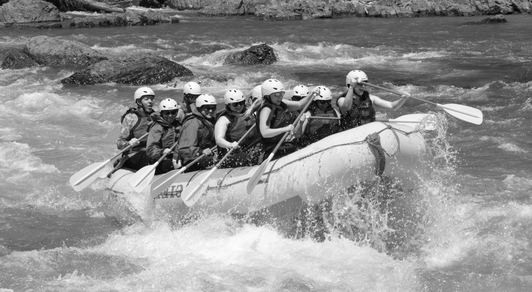 Abgebildet sind Frauen und Männer in einem Rafting-Boot, die gemeinsam das Boot durch bewegtes Wasser steuern. Die Teammitglieder lachen.