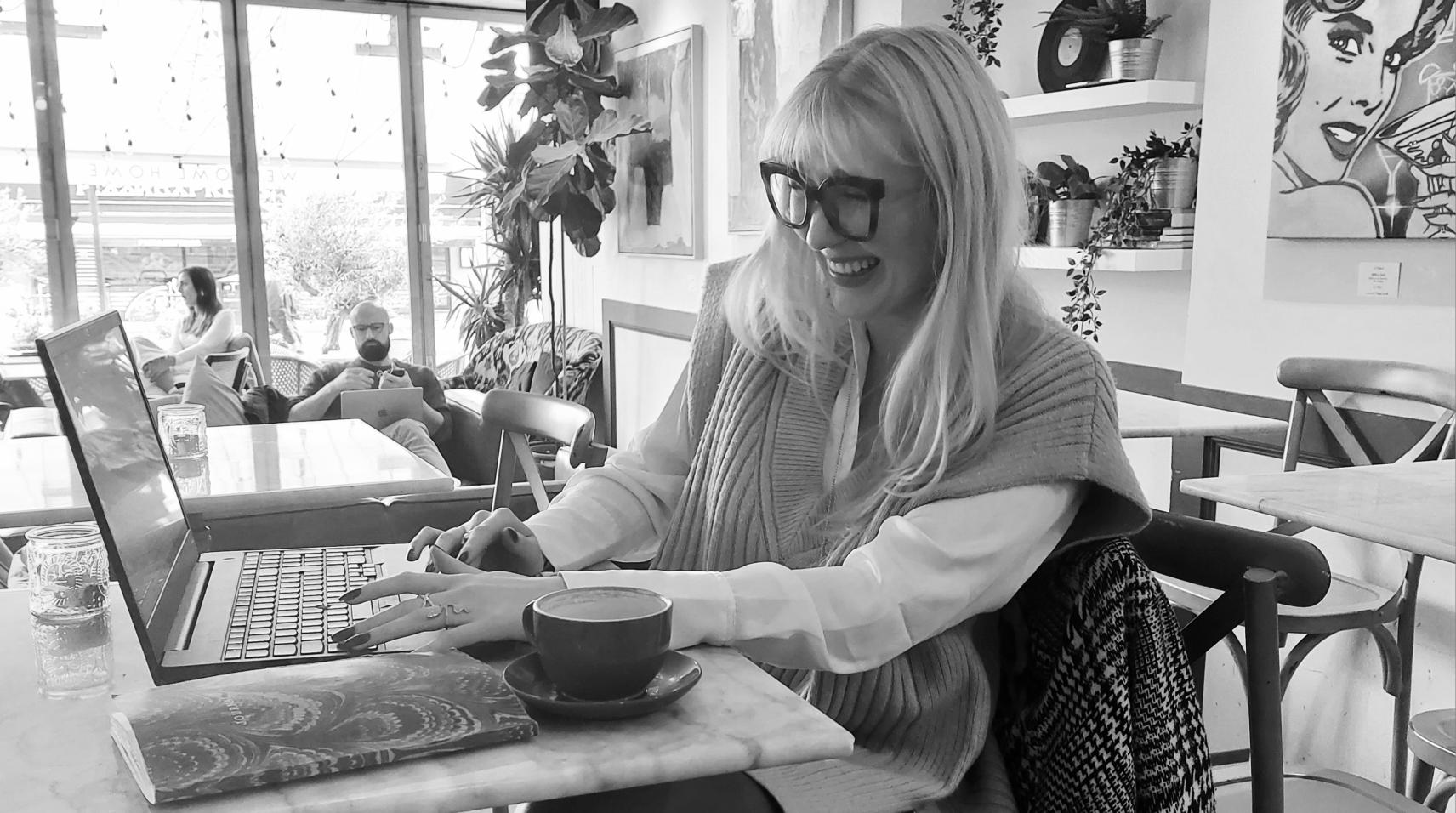 Alexandra van Hall sitzt in einem Café an einem Tisch vor ihrem Laptop. Sie tippt auf der Tastatur und lacht, während ihr Blick auf der Bildschirm weilt.