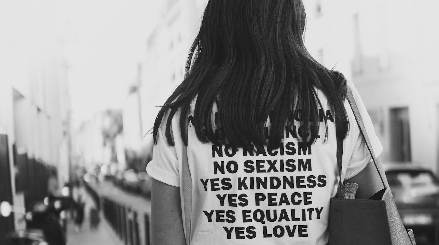 Bild von einer jungen Frau, deren Rücken nur zu erkennen ist. Auf ihrem T-Shirt sind die Worte "No racism", "No "sexism", "Yes Kindness", "Yes Peace", "Yes Equality", "Yes Love" zu lesen.
